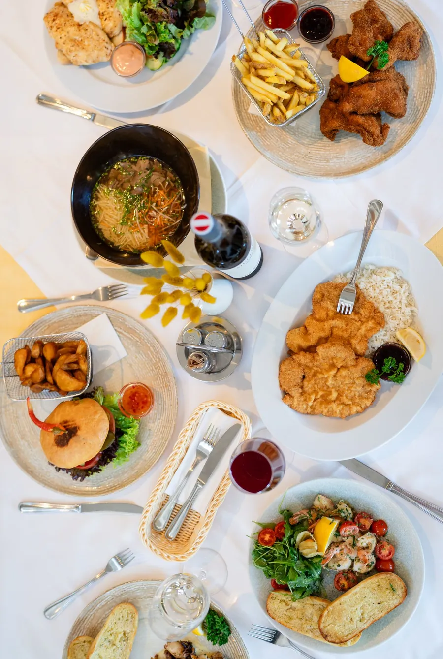 Gedeckter Tisch von oben mit Backhendl, Schnitzel, Suppe, Burger mit Erdäpfelspalten, Garnelen, Wein und Brotkorb